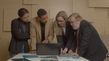 Professional successful diverse multiracial business team gathering around laptop, discussing financial data, sharing ideas and visions while working and planning company strategy in boardroom.