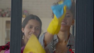 Portrait of happy cute multiethnic teen daughter with pigtails and positive attractive African American mother doing spring cleanup, cleaning window glass using spray detergent and rags.