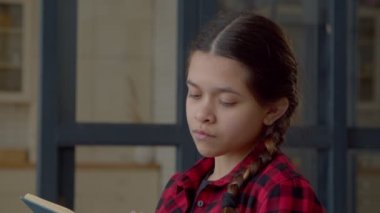 Portrait of thoughtful engrossed in reading attractive Latin teen girl with active imagination and stylish pigtails spending pastime, enjoying imaginative literature while relaxing indoors.