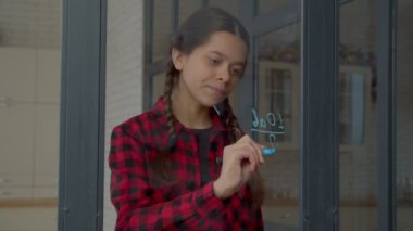 Thoughtful clever lovely Hispanic adolescent schoolgirl with pigtails studying homework, writing with felt tip pen and solving high school mathematical example on window glass indoors.