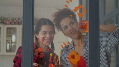 Joyful lovely teen daughter with pigtails and cheerful charming African American mother enjoying pastime, and having fun , painting with hands on window glass using colorful paints.