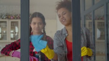 Positive adorable multicultural teenage daughter with pigtails and cheerful pretty African American mother in rubber gloves doing housework together , cleaning window using spray detergent and rags.