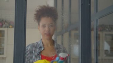 Positive pretty African American woman in rubber gloves doing domestic chores, washing window glass using spray detergent and rag during spring cleaning at home.