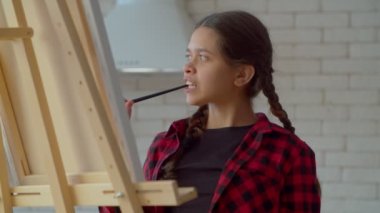 Portrait of pensive lovely Latin teen girl artist with paintbrush in mouth thinking about ideas for painting, finding inspiration and creative impulse for artwork while spending leisure at home.