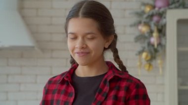 Portrait of positive adorable Hispanic teenager girl with pigtails applying eye black underneath eyes, looking with radiant smile, showing carefree mood, happiness and joy indoors.