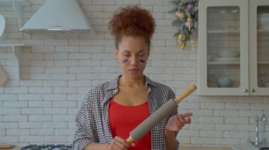 Portrait of strict determined attractive African American female wearing eye black, holding rolling pin and making aggressive gesture, showing irritation , fury and anger indoors.