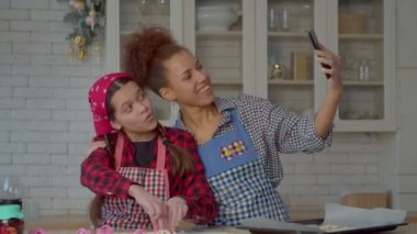 Excited playful multicultural teen daughter and joyful pretty African American mother having fun, posing for selfie shot on smartphone, making funny faces and gestures during cooking in kitchen.