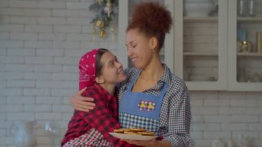 Çekici siyahi anne portresi ve önlüklü çok ırklı mutlu genç kız elinde ev yapımı kurabiyelerle, sevgiyle kucaklaşarak, mutfakta mutluluk ve neşe ifade ederek.