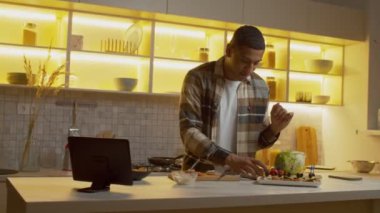 Handsome black male preparing tasty canapes with fresh salmon and blue cheese, decorating snack with fresh olive and slice of cucumber on stick while cooking dinner in domestic kitchen.
