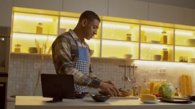 Positive handsome black male cook in apron slicing baked chicken fillet on cutting board, preparing food ingredients for delicious fresh salad while cooking dinner in domestic kitchen.