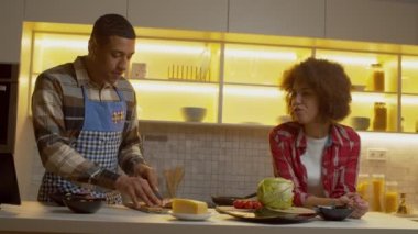 Handsome African American man in apron preparing romantic dinner in domestic kitchen, forbidding hungry charming black woman to taste slice of baked chicken breast while cooking in kitchen.