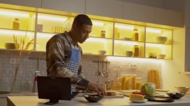 Strict engrossed in cooking attractive African American man in apron cooking dinner, chopping food ingredients on cutting board and ,refusing help from pretty black woman, in domestic kitchen.