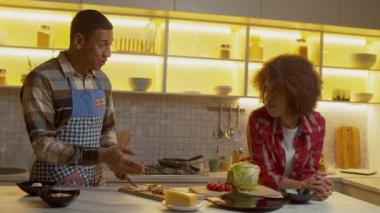 Charming positive African American woman tasting slice of delicious baked chicken cooked by loving black man, while husband in apron chopping meat on cutting board during cooking in kitchen.