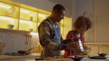 Joyful handsome African American man in apron grating cheese on grater and positive lovely black woman slicing cabbage, while happy couple preparing fresh delicious salad together in kitchen
