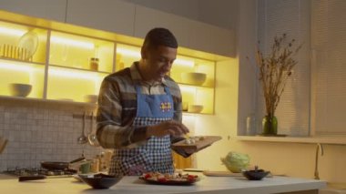 Positive handsome African American man in apron placing sliced quail eggs into delicious fresh salad from cutting board, while preparing tasty course during cooking romantic dinner in kitchen