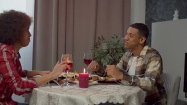 Affectionate handsome African American man and pretty black woman enjoying romantic dinner and bonding, toasting with red wine glasses while celebrating event in domestic room.