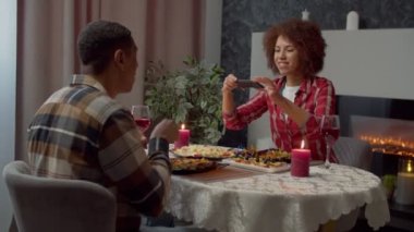 Cheerful lovely African American woman taking food selfie on smartphone and posting in social media, while happy affectionate couple enjoying romantic dinner in domestic room.