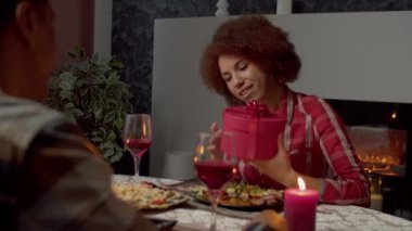 Portrait of charming excited black female shaking gift box, trying to guess present, given by affectionate African American man on valentines day while couple relaxing at festive table.