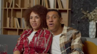 Portrait of attractive positive black spouses sipping red wine from glasses, watching interesting movie on tv and chatting while enjoying leisure together inn domestic room.