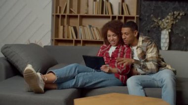 Positive attractive African American couple sitting on couch in love embrace, sharing digital tablet, networking and discussing online social media content while enjoying leisure at home.