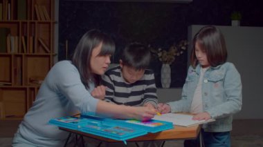 Positive attractive Asian mother and adorable preadolescent children developing creative skills and artistic imagination, designing picture on paper sheet with colorful felt tip pens in domestic room.