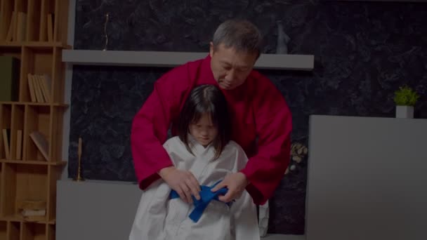 英俊的资深跆拳道大师帮助可爱的少年前武术亚洲女学生为训练准备校服 在室内用扁结系上蓝带 — 图库视频影像