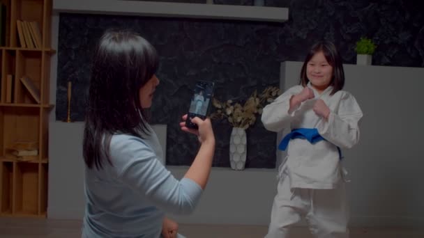 快乐可爱的前亚洲跆拳道少女 身穿制服 摆出一副打斗姿势 表现出武术技巧 而快乐的母亲则在室内用手机给女儿拍照 — 图库视频影像