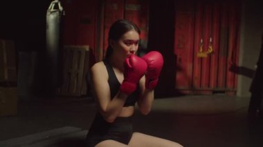 Motivasyonlu spor boks eldivenli çekici Asyalı kadın antrenmana hazırlanıyor, boks kombinasyonları yapıyor ve grunge spor salonunda paletlerin üzerinde otururken yumruk tekniklerini geliştiriyor..