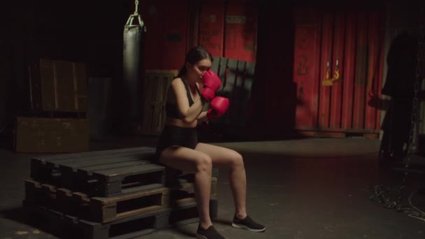 在黑暗的车库体育馆里 穿着手套准备运动训练的亚洲女拳击手坐在货盘上 练习拳击技术 并决定了严肃而有吸引力的运动健将 — 图库视频影像