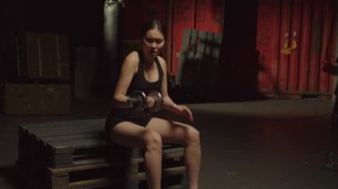 Aktif konsantrasyonlu spor kıyafetleri içinde güzel bir Asyalı kadın spor yaparken omuz kaslarını sakatlıyor halterle ağırlık çalışırken koluna acı dolu bir ifadeyle dokunuyor ve karanlık garajda spor yaparken spor yapıyor..