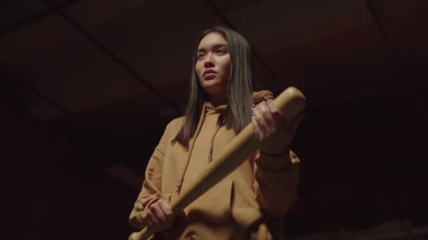 在黑暗的废弃建筑里 顽强而坚定的 漂亮的亚裔女性罪犯手握棒球棒 凝视着粗糙而集中的眼神 表现出威胁和挑衅的低视角 — 图库视频影像