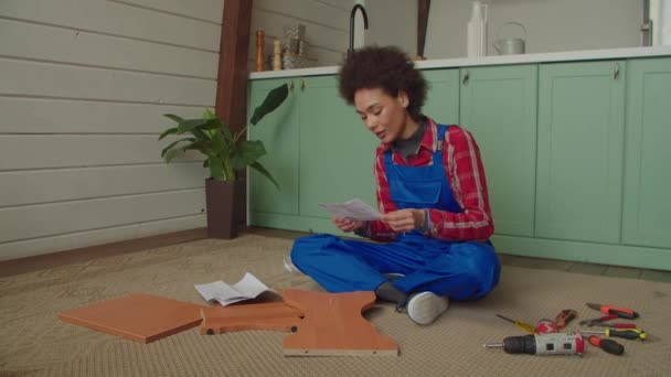全神贯注的非洲裔美国年轻女性 身穿防护服 双腿交叉地坐在地板上 拿着工作和电动工具 阅读在家里组装Diy家具的说明 — 图库视频影像