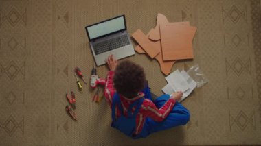 Koruyucu iş kıyafetleri ve elektrikli aletleri olan çekici siyahi kadınların en iyi görüntüsü yerde oturmuş, bilgisayardan video dersleri izliyor, DIY mobilyalarını evde monte etmeyi öğreniyor..