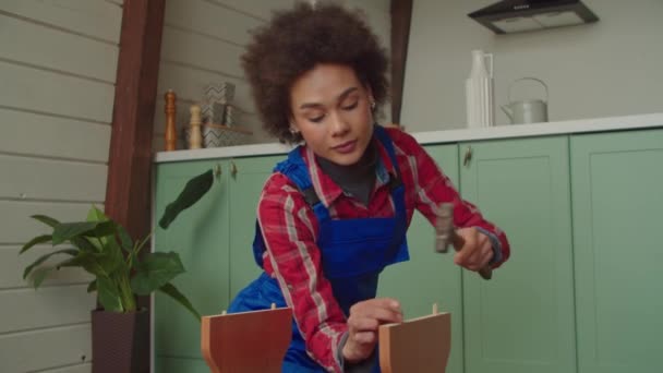 作業服に集中魅力的な黒の女性は 国内の部屋で作業ツールを使用してDiy家具を組み立てながら 人間と木製のタオルピンを爪 スツールシートを調整します — ストック動画