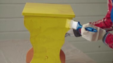 Koruyucu eldivenli kadın ellerinin yakın çekimi boya fırçasıyla yeniden kullanım için eski mobilyaları restore etmek, sarı renkte tabureyi boyamak, ev işi yapan kadın kapalı alanda çalışırken.