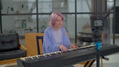 Neşeli yetenekli çekici bayan müzisyen dijital kamerayla sosyal medya için vlog kaydediyor, piyano klavyesi çalıyor ve evde boş vakitlerin tadını çıkarırken online seyirciler için yeni bir şarkı söylüyor..
