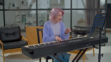 Pozitif yetenekli pembe saçlı bayan müzisyen elektronik piyano klavyesi çalıyor, şarkı yazarken yakalama şarkısı söylüyor yerli odada hit oldu..
