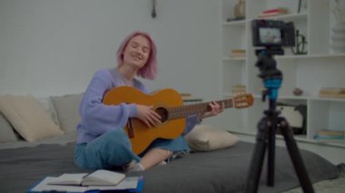 Çekici pembe saçlı kadın söz yazarı sosyal medya için kamera önünde video içeriği kaydediyor, akustik gitar çalıyor ve yatak odasında boş vaktini geçirirken yeni bir şarkı söylüyor..