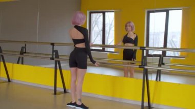 Dikiz aynasından yansıyan aynaya bakarak dans stüdyosunda prova yaparken Demi Plie egzersizi yapan zarif, yoğun pembe saçlı kadın balerin..