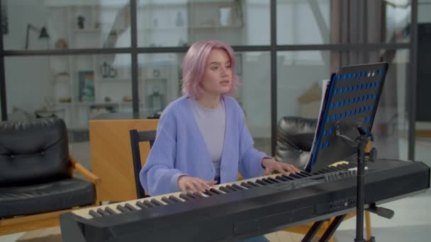 美しいピンク色の髪の女性のピアニストが演奏し 電子ピアノのキーボードを演奏し 国内の部屋で練習しながら シートミュージックスタンドに音楽ノートでページを残します — ストック動画