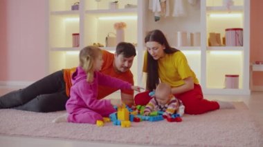 Sevecen, çekici ebeveynler, neşeli küçük bir kız çocuğu ve neşeli bir anaokulu kızı birlikte eğleniyorlar, renkli çocuklarla oynuyorlar, evcil odaların zemininde taş inşa ediyorlar..
