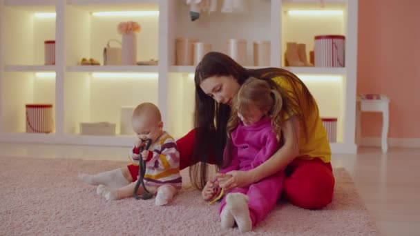 有爱心的积极而有魅力的母亲 有可爱的女婴和可爱的学龄前女儿坐在地板上 在家里放松的同时 与五彩缤纷的玩具相处和玩耍 — 图库视频影像