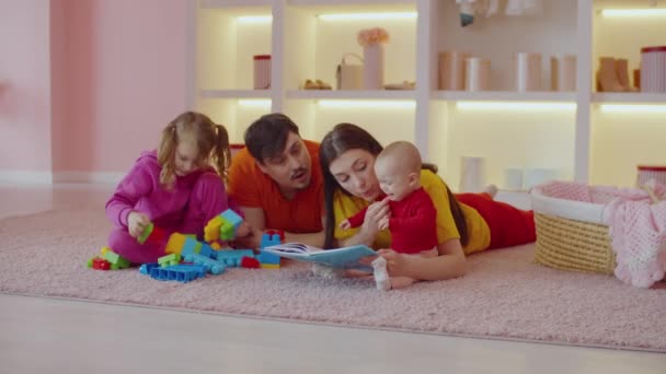 慈祥英俊的父亲和兴奋的可爱小女儿在地板上玩玩具块 而可爱迷人的母亲和心烦意乱的可爱婴儿在客厅里读童话 — 图库视频影像