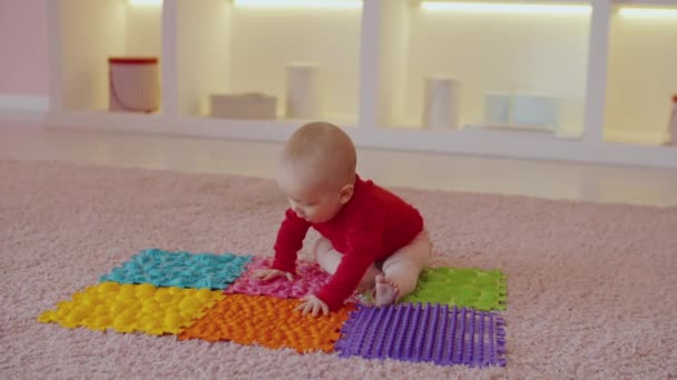 カラフルな整形外科マッサージパズルマットに座って遊んでいる好奇心かわいい裸足の幼児は 屋内で肯定的で陽気な気分を表現し 新しいものの発見への関心を示しています — ストック動画