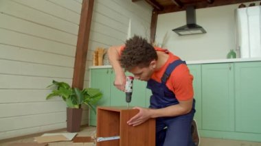 Koruyucu iş giysisinde yetenekli Afrikalı Amerikalı adam DIY komodin montajı, iç odada çalışırken elektrikli matkap tornavidayla vidaları sıkma.