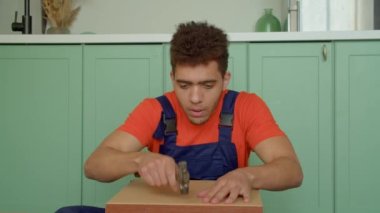 Koruyucu iş giysisinde yetenekli siyah erkek boyanın portresi yere oturmuş, komodinin arka tarafına çekiçle çivi çakıyor ve evde DIY mobilya monte ediyor..