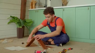 Koruyucu iş giysisinde konsantre olmuş yakışıklı Afrikalı Amerikalı tamirci komodinin çekmecesinin bir kısmını tamir ediyor, evde DIY mobilya montajı sırasında iş aletini kullanarak vidaları sıkıyor..
