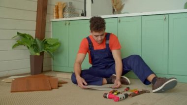 Konsantre olmuş, koruyucu iş kıyafetleri içinde, iş ve elektrikli aletlerle yerde oturan ve DIY mobilyalarını evde monte etmek için talimat okuyan Afro-Amerikan genç adam..