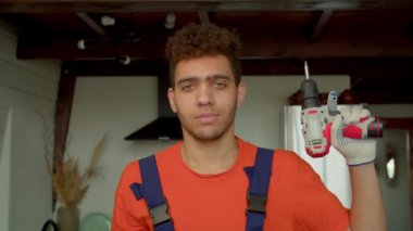 Koruyucu iş giysisinde, elinde kablosuz elektrikli matkap tornavidayla, kendine güvenen, yakışıklı bir erkek boksör. Baş parmak hareketi yapıyor ve yurt içi odasında neşeli bir gülümsemeyle bize bakıyor..