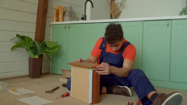 Koruyucu iş elbisesi içinde konsantre olmuş yakışıklı siyah tamirci DIY komodin çekmecesinin montajını tamamlıyor, oturma odasında yerde otururken ön tahta paneli ayarlıyor..